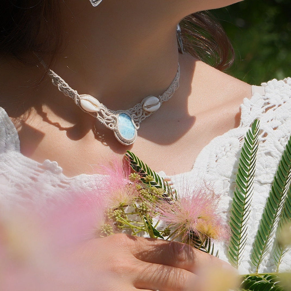 Collier plastron en Larimar Océan bleu ciel, coton couleur lin naturel (beige gris), cauris blancs et perles en acier inoxydable argenté. Système de réglable en tube tissé à l'arrière du bijou. Tissage symétrique en micro macramé avec effet nœuds celtiques entrelacés en relief. Style bohème, hippie, plage, Sirène, tribal et tropical. Convient pour un mariage type bohème ou plage. Bijou porté avec une tenue romantique et bohème blanche avec dentelle apparente. Modèle à la peau bronzée et aux cheveux bruns. 