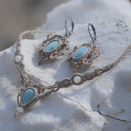 Parure Trésors d'Ecume composée d'une paire de boucles d'oreilles pendentes avec collier ras de cou assorti. Larimar bleu ciel ovales pour les boucles et en poire pointe en bas pour le collier. 3 Perles en nacre blanche pour le collier. Perles acier inoxydable argenté pour les deux pour le côté luxe et précieux. Dormeuses en acier inoxydable pour un port assuré. Fil de Coton couleur lin naturel (beige gris) de 0, 5 mm tissé en micro-macramé en relief version celtique. Bijoux posés sur une pierre blanche 