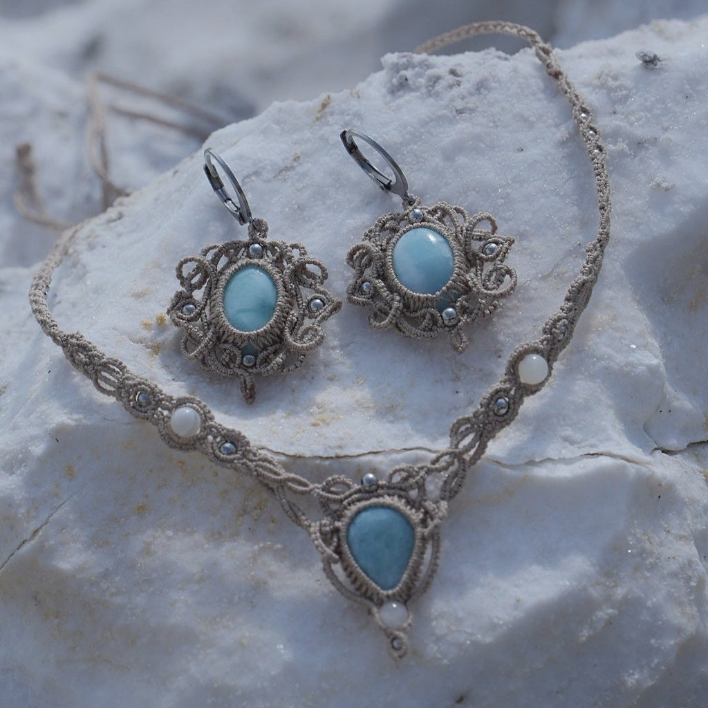 Parure Trésors d'Ecume composée d'une paire de boucles d'oreilles pendentes avec collier ras de cou assorti. Larimar bleu ciel ovales pour les boucles et en poire pointe en bas pour le collier. 3 Perles en nacre blanche pour le collier. Perles acier inoxydable argenté pour les deux pour le côté luxe et précieux. Dormeuses en acier inoxydable pour un port assuré. Fil de Coton couleur lin naturel (beige gris) de 0, 5 mm tissé en micro-macramé en relief version celtique. Bijoux posés sur une pierre blanche 