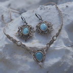 Parure Trésors d'Ecume composée d'une paire de boucles d'oreilles pendentes avec collier ras de cou assorti. Larimar bleu ciel ovales pour les boucles et en poire pointe en bas pour le collier. 3 Perles en nacre blanche pour le collier. Perles acier inoxydable argenté pour les deux pour le côté luxe et précieux. Dormeuses en acier inoxydable pour un port assuré. Fil de Coton couleur lin naturel (beige gris) de 0, 5 mm tissé en micro-macramé en relief version celtique. Bijoux posés sur une pierre blanche 