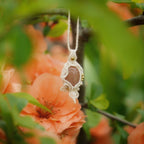 Collier pendentif en micro macramé avec pierre de soleil pêche et nacre, posé sur une branche de cognassier en fleurs saumonées, style bohème et féérique.