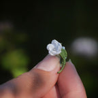 Broche miniature florale blanche en micro macramé avec labradorite bleu-vert au centre, tenue dans une main sur fond de feuilles vertes, bijou bohème et elfique artisanal.
