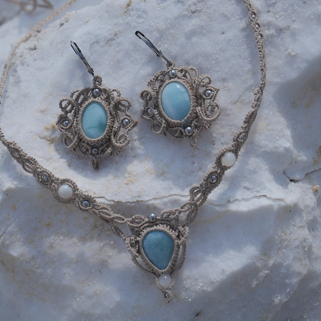 Parure Trésors d'Ecume composée d'une paire de boucles d'oreilles pendentes avec collier ras de cou assorti. Larimar bleu ciel ovales pour les boucles et en poire pointe en bas pour le collier. 3 Perles en nacre blanche pour le collier. Perles acier inoxydable argenté pour les deux pour le côté luxe et précieux. Dormeuses en acier inoxydable pour un port assuré. Fil de Coton couleur lin naturel (beige gris) de 0, 5 mm tissé en micro-macramé en relief version celtique. Bijoux posés sur une pierre blanche 