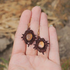 « Boucles d’oreilles artisanales elfique en citrine et grenat, bijou bohème léger et raffiné tissé en micro macramé, présenté dans une main à peau claire »