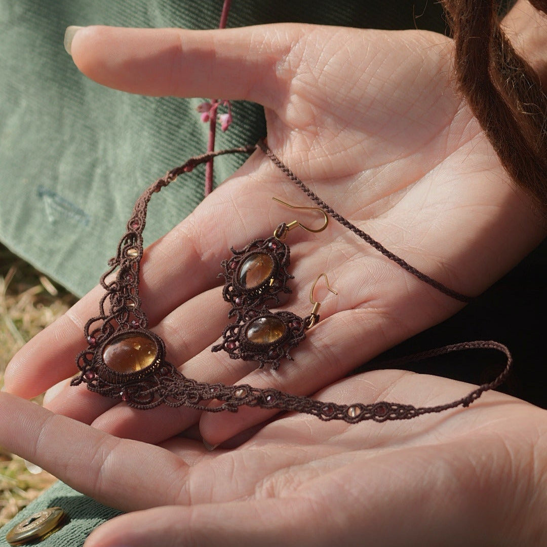 Parure elfique artisanale en micro macramé brun, composée d’un collier ras de cou réglable en citrine jaune et grenat rouge rosé, assorti à des boucles d’oreilles florales. Ensemble bohème et féérique de la collection Clairière de Miel, présenté dans une main féminine à la peau claire.