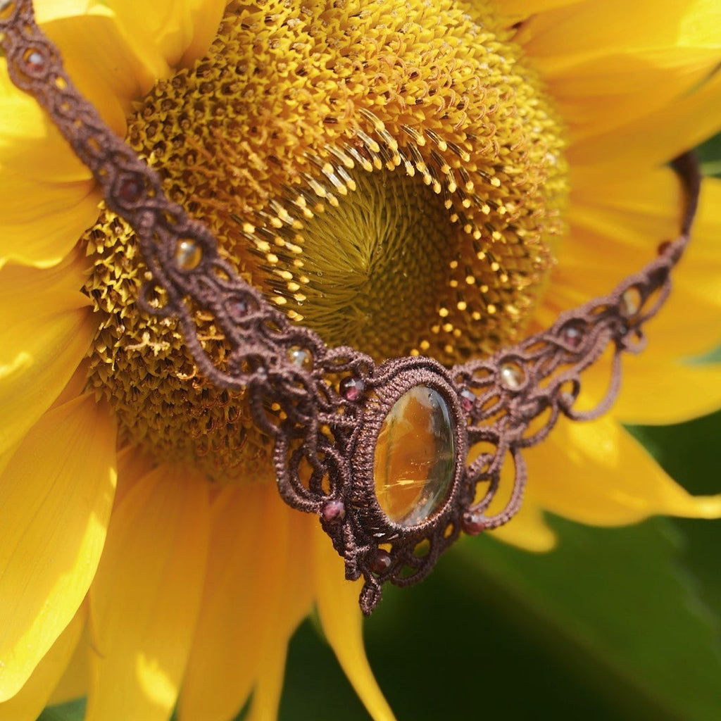 Collier ras de cou elfique en citrine et grenat, fait main en micro macramé brun. Bijou bohème et solaire de la collection Clairière de Miel, photographié dans la nature parmi des tournesols rappelant la couleur dorée de la citrine.