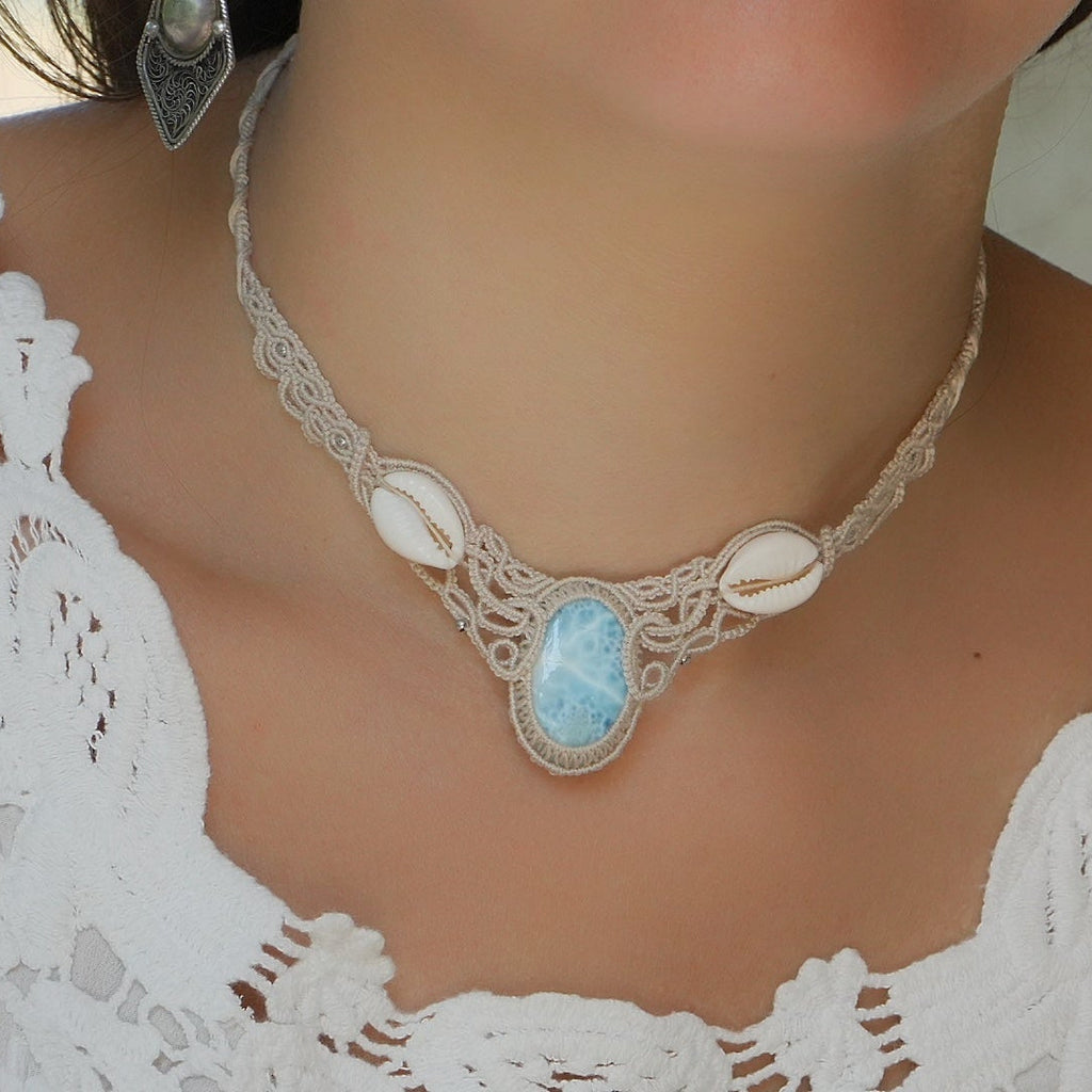 Collier plastron en Larimar Océan bleu ciel, coton couleur lin naturel (beige gris), cauris blancs et perles en acier inoxydable argenté. Système de réglable en tube tissé à l'arrière du bijou. Tissage symétrique en micro macramé avec effet nœuds celtiques entrelacés en relief. Style bohème, hippie, plage, Sirène, tribal et tropical. Convient pour un mariage type bohème ou plage. Bijou porté avec une tenue romantique et bohème blanche avec dentelle apparente. Modèle à la peau bronzée et aux cheveux bruns. 
