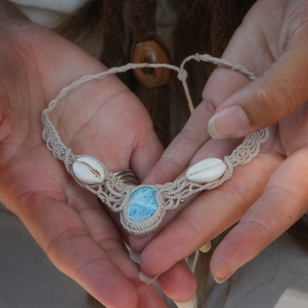 Collier plastron en Larimar Océan bleu ciel, coton couleur lin naturel (beige gris), cauris blancs et perles en acier inoxydable argenté. Système de réglable en tube tissé à l'arrière du bijou. Tissage symétrique en micro macramé avec effet nœuds celtiques entrelacés en relief. Style bohème, hippie, plage, Sirène, tribal et tropical. Convient pour un mariage type bohème ou plage. Bijou tenu vue 3/4, tenue romantique et bohème blanche avec dentelle apparente. Modèle à la peau bronzée et aux cheveux bruns. 