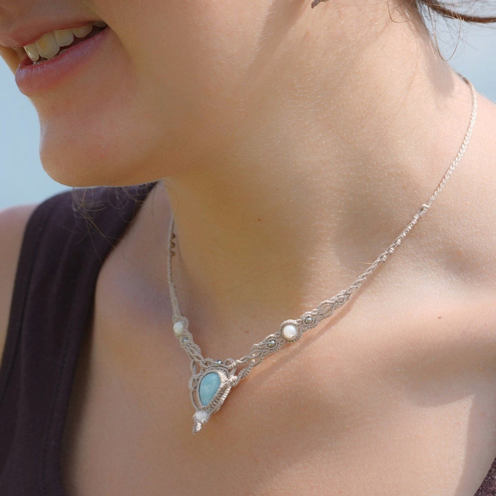 Collier ras-de-cou forme v Trésors d'Ecume composée d'une Larimar bleu ciel en poire pointe en bas, perles en nacre blanche ronde rappel des coquillages et de la plage, mini Perles rondes en acier inoxydable argenté pour le côté luxe et précieux. Fil de Coton couleur lin naturel (beige gris) de 0, 5 mm tissé en micro-macramé en relief version celtique. Collage 3 photos : Bijoux porté sur une femme bronzée et brune avec débardeur marron foncé. Photo ambiance tropiques et plages, style bohème hippie 