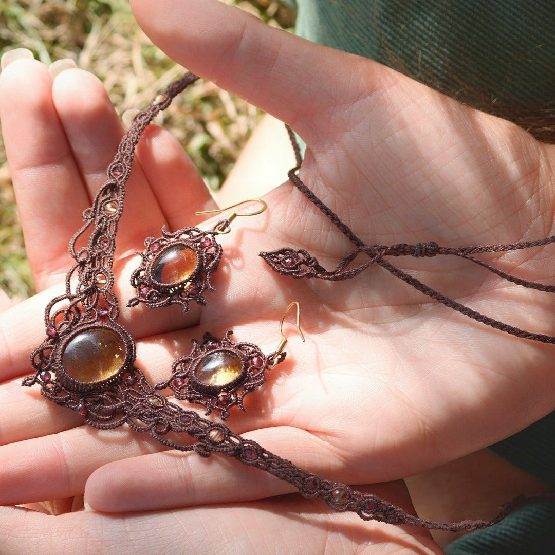 Parure elfique artisanale en micro macramé brun, composée d’un collier ras de cou réglable en citrine jaune et grenat rouge rosé, assorti à des boucles d’oreilles florales. Ensemble bohème et féérique de la collection Clairière de Miel, présenté dans une main féminine à la peau claire.