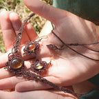 Parure elfique artisanale en micro macramé brun, composée d’un collier ras de cou réglable en citrine jaune et grenat rouge rosé, assorti à des boucles d’oreilles florales. Ensemble bohème et féérique de la collection Clairière de Miel, présenté dans une main féminine à la peau claire.