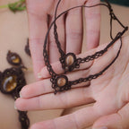 Bijoux de pieds, paire assortie de bracelets de chevilles à ambiance bohème celtique elfique. Fil marron foncé fin tissé en micro macramé avec entrelacs et arabesques, pierre centrale ovale petite taille jaune miel translucide. Bijoux hippie, bohème avec inspiration nature. Chevillères posée à plat dans 2 mains en coupe l'une au dessus de l'autre avec en fond d'autres bijoux dans les mêmes tons en citrine