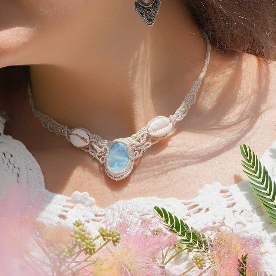Collier plastron en Larimar Océan bleu ciel, coton couleur lin naturel (beige gris), cauris blancs et perles en acier inoxydable argenté. Système de réglable en tube tissé à l'arrière du bijou. Tissage symétrique en micro macramé avec effet nœuds celtiques entrelacés en relief. Style bohème, hippie, plage, Sirène, tribal et tropical. Convient pour un mariage à thème et festival. Bijou porté avec une tenue romantique et bohème blanche avec dentelle apparente. Modèle à la peau bronzée et aux cheveux bruns. 