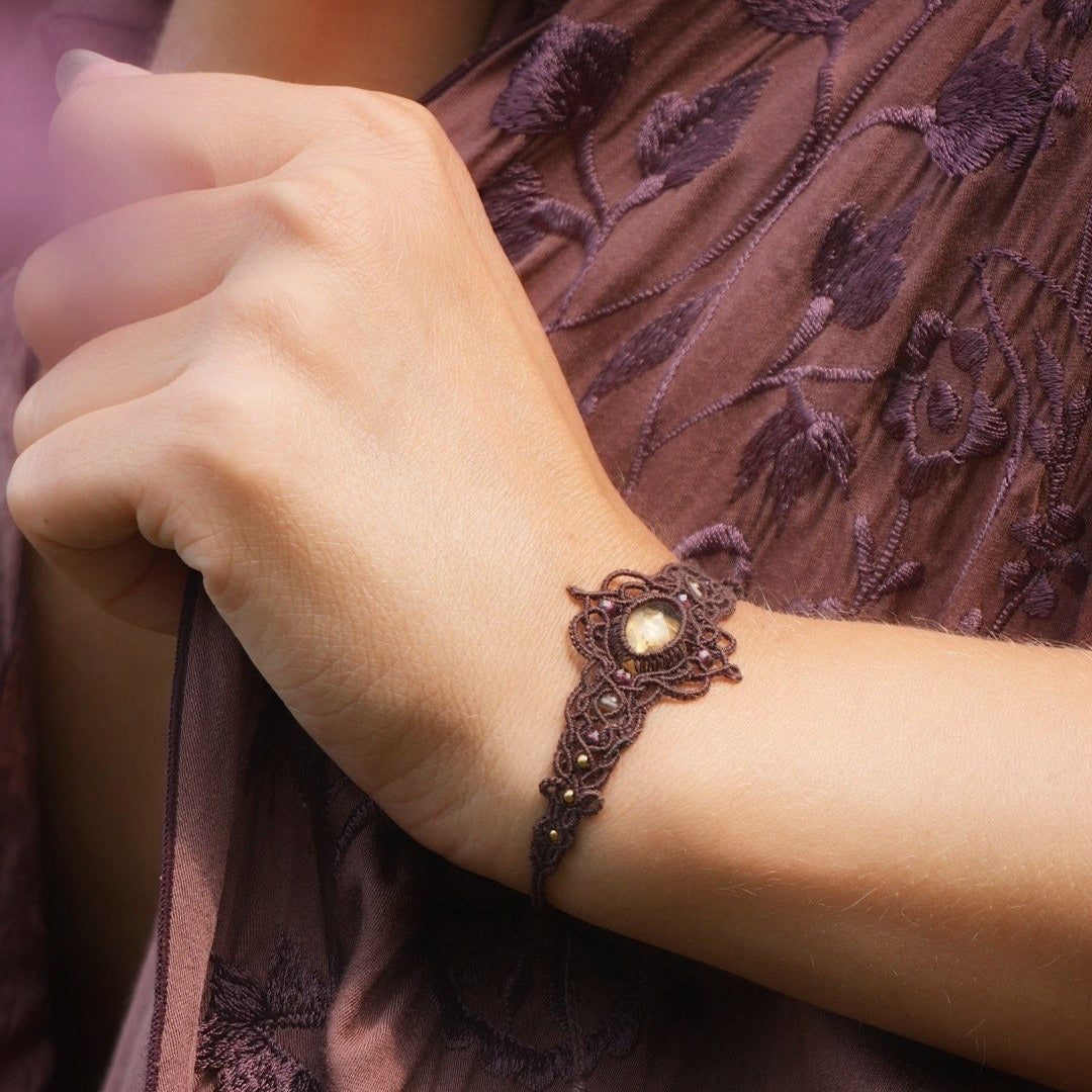 Bracelet large elfique en micro macramé brun, orné d’une citrine jaune pâle et de perles de grenat rouge rosé. Bijou bohème et artisanal réglable, porté au poignet d’une femme à la peau claire.
