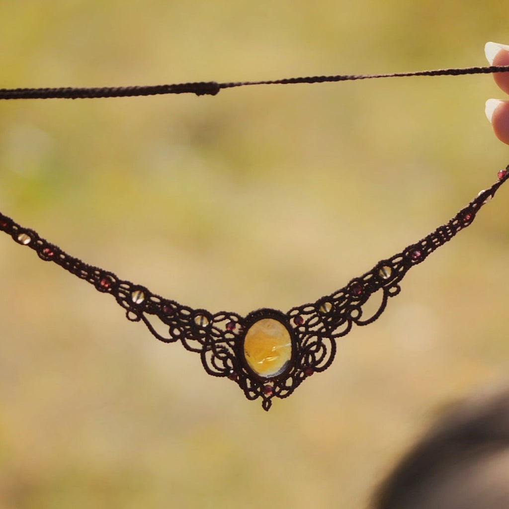 Collier elfique en macramé fait main, avec citrine ovale translucide jaune ambré et perles de grenat rouge rosé. Bijou artisanal de la collection Clairière de Miel, présenté en main pour révéler la transparence lumineuse de la pierre.