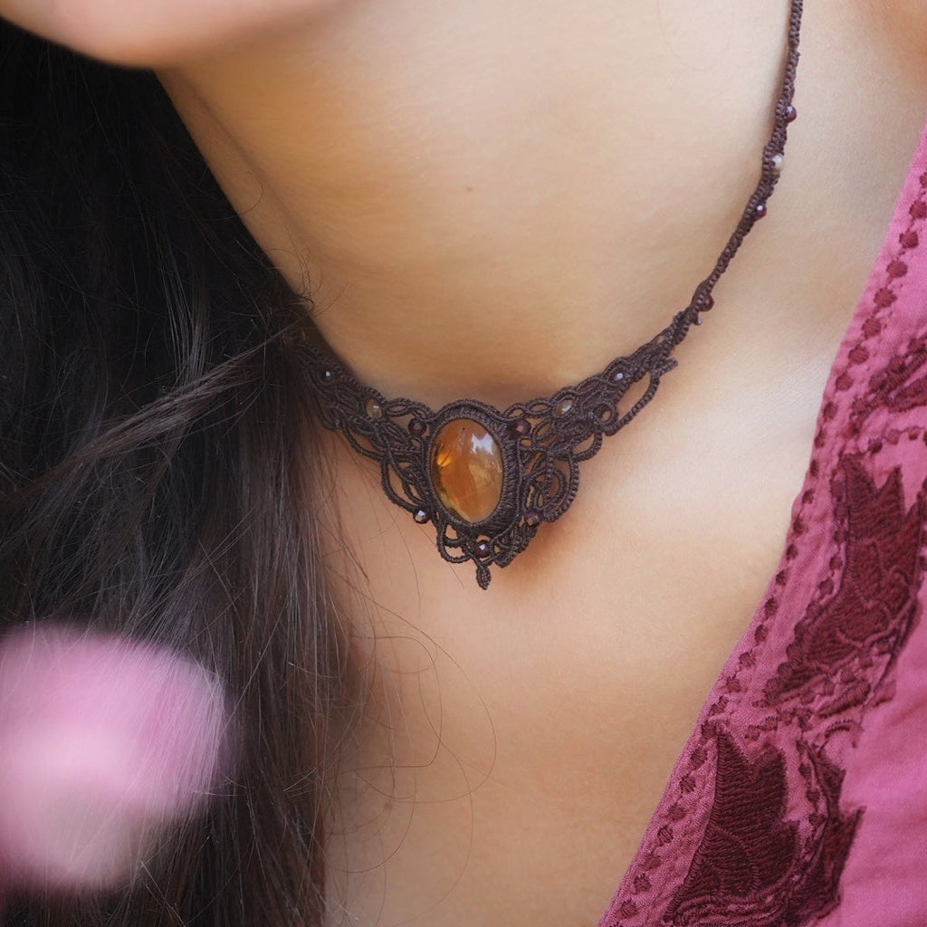 Collier ras de cou elfique en micro macramé brun, orné de citrine jaune dorée et de grenat rouge rosé. Bijou bohème réglable, porté par une femme à la peau claire et cheveux bruns, assorti à un vêtement rose profond.