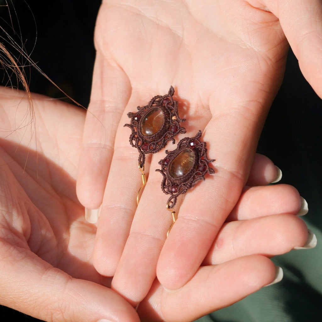 « Boucles d’oreilles artisanales elfique en citrine et grenat, bijou bohème léger et raffiné tissé en micro macramé, présenté dans une main à peau claire »