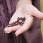 Bracelet elfique artisanal en citrine et grenat, réglable et tissé à la main en micro macramé brun. Bijou unique et lumineux, présenté dans une main féminine à la peau claire.