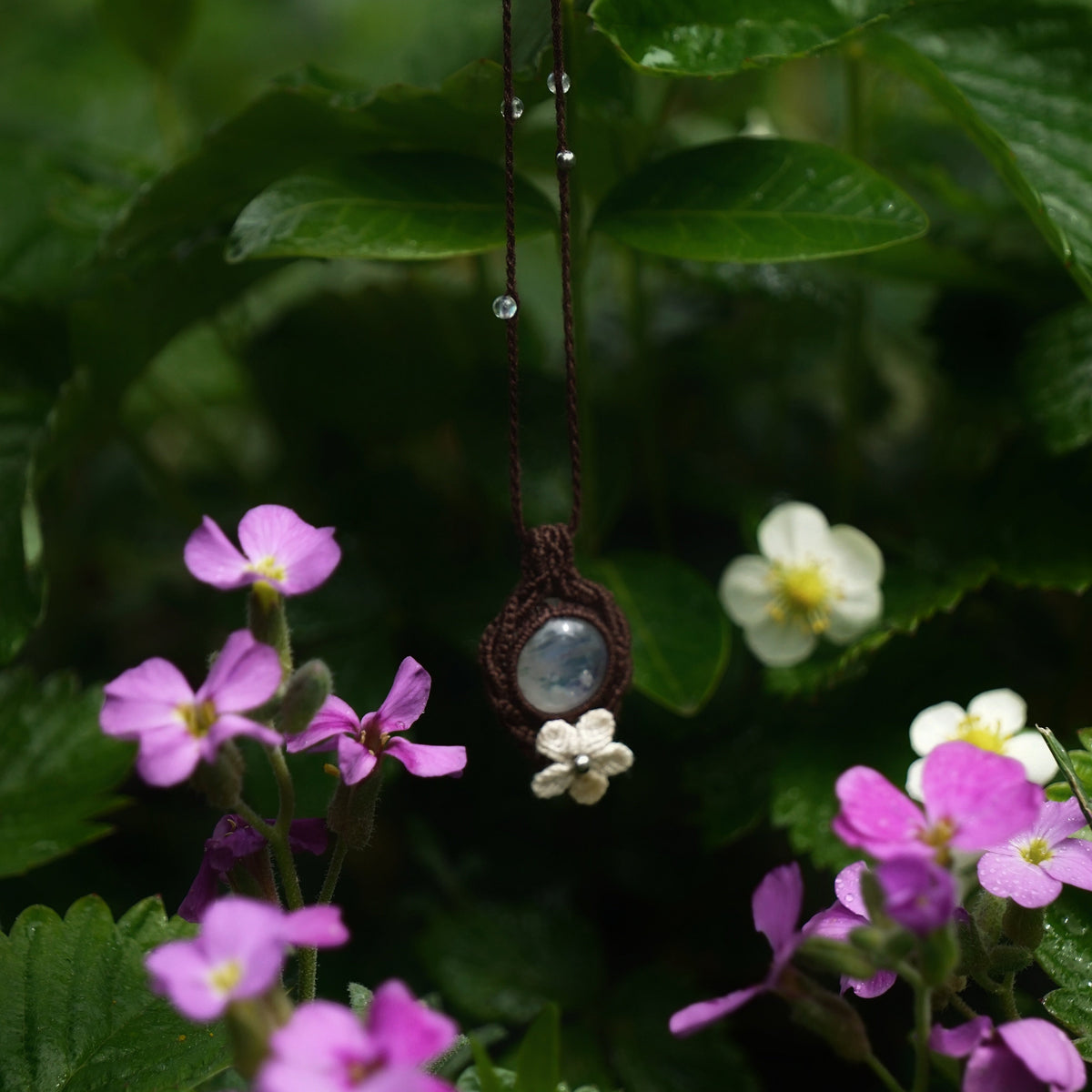 Bijou pendentif en labradorite blanche suspendu verticalement au milieu de fleurs roses et de feuilles de fraisiers mouillées par la pluie, collier elfique bohème réglable. Coton et Astrance