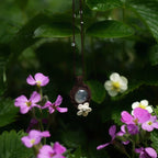Bijou pendentif en labradorite blanche suspendu verticalement au milieu de fleurs roses et de feuilles de fraisiers mouillées par la pluie, collier elfique bohème réglable. Coton et Astrance