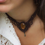 Collier large en micro macramé de couleur marron foncé, jaune ambré et bronze. Motif tissé en micro macramé avec ambiance entrelacs celtiques et arabesques elfiques. Présence d'une pierre centrale ovale de grande taille jaune translucide, gouttes bronze, forme V avec mini perles ronde bronze parsemées. Photo ambiance bohème hippie avec une touche d'élégance. Bijou porté sur une peau claire avec chemise blanche en dentelle.
