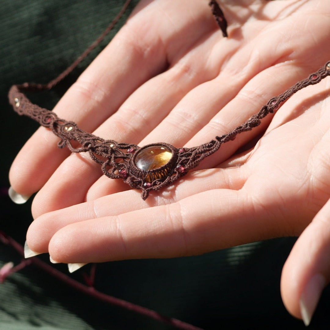 Bijou elfique artisanal réglable en citrine jaune et grenat rouge, tissé en macramé brun. Collier ras de cou bohème, présenté dans une main féminine à la peau claire, mettant en valeur la finesse du tissage.
