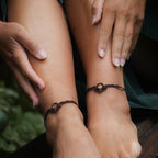 Bijoux de pieds, paire assortie de bracelets de chevilles à ambiance bohème celtique elfique. Fil marron foncé fin tissé en micro macramé avec entrelacs et arabesques, pierre centrale ovale petite taille jaune miel translucide. Bijoux hippie, bohème avec inspiration nature. Chevillères portées sur des chevilles à la peau pâle, ambiance sombre/dark. Modèle assise sur un banc en bois, les jambes relevées, les mains autour des mollets, de face avec jeu d'ombre en fond, jupe velours côtelé vert sapin