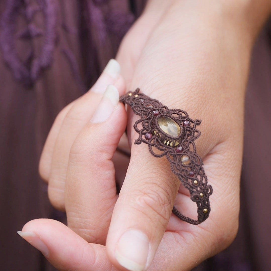 Bracelet elfique artisanal en citrine et grenat, réglable et tissé à la main en micro macramé brun. Bijou unique et lumineux, présenté dans une main féminine à la peau claire.