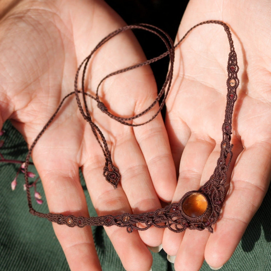 Bijou elfique artisanal réglable en citrine jaune et grenat rouge, tissé en macramé brun. Collier ras de cou bohème, présenté dans une main féminine à la peau claire, mettant en valeur la finesse du tissage.