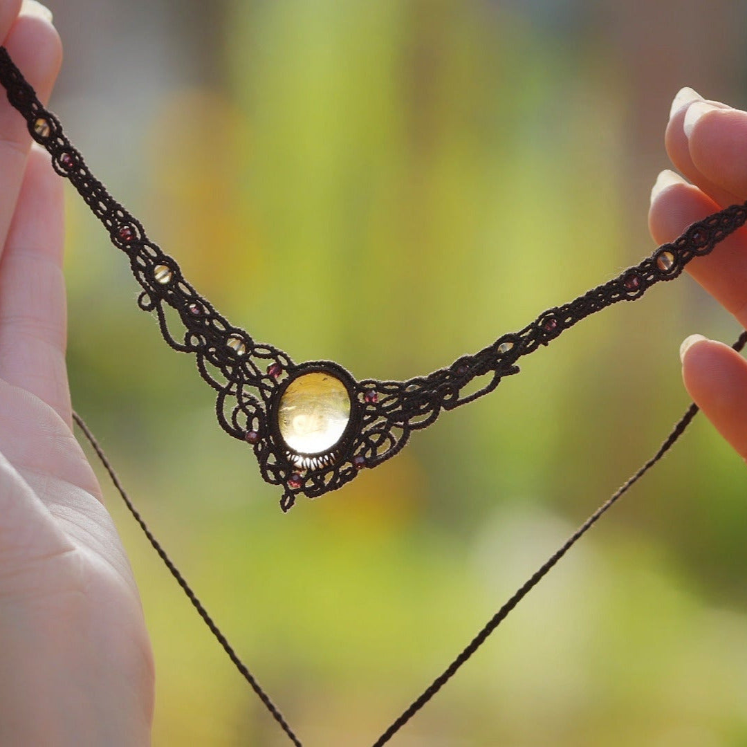 Collier elfique en macramé fait main, avec citrine ovale translucide jaune ambré et perles de grenat rouge rosé. Bijou artisanal de la collection Clairière de Miel, présenté en main pour révéler la transparence lumineuse de la pierre.