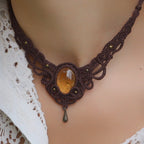 Collier large en micro macramé de couleur marron foncé, jaune ambré et bronze. Motif tissé en micro macramé avec ambiance entrelacs celtiques et arabesques elfiques. Présence d'une pierre centrale ovale de grande taille jaune translucide, gouttes bronze, forme V avec mini perles ronde bronze parsemées. Photo ambiance bohème hippie avec une touche d'élégance. Bijou porté sur une peau claire et chemise blanche en dentelle.