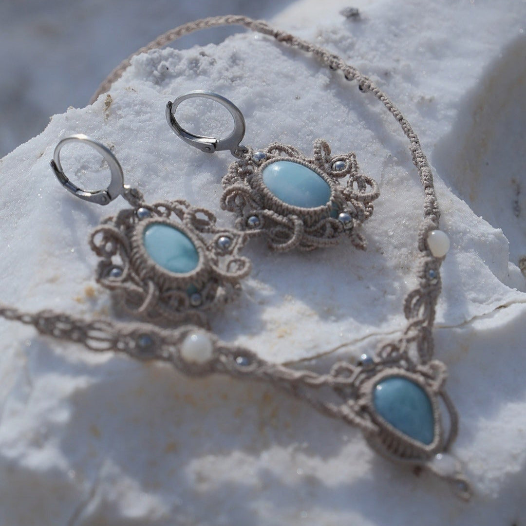 Parure Trésors d'Ecume composée d'une paire de boucles d'oreilles pendentes avec collier ras de cou assorti. Larimar bleu ciel ovales pour les boucles et en poire pointe en bas pour le collier. 3 Perles en nacre blanche pour le collier. Perles acier inoxydable argenté pour les deux pour le côté luxe et précieux. Dormeuses en acier inoxydable pour un port assuré. Fil de Coton couleur lin naturel (beige gris) de 0, 5 mm tissé en micro-macramé en relief version celtique. Bijoux posés sur une pierre blanche 