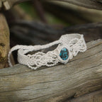Bracelet large asymétrique. Bijou dentelle ambiance bohème hippie. Bracelet tissé en micro macramé avec fil de coton écru de 0.5 mm avec une pierre ovale de chrysocolle couleur bleu turquoise et marron foncé chocolat. Présence de part et d'autre de façon symétrique des perles en acier inoxydable argentées. Système de réglage à coulisse tubulaire à l'arrière. Design en relief et en torsade entrelacées celtiques. Ambiance plages, festival d'été et mariage boho. Bijou posé sur un galet beige et coquillages.