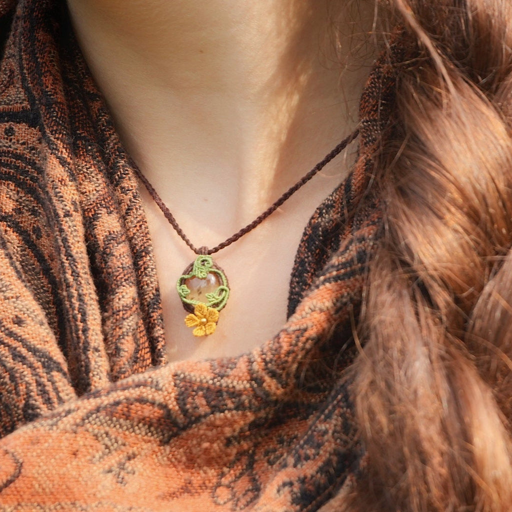 Collier elfique réglable en micro macramé avec citrine ovale jaune pâle et fleur orange dorée. Bijou floral et lumineux de la collection Clairière de Miel, porté par une femme à la peau claire et cheveux bruns.