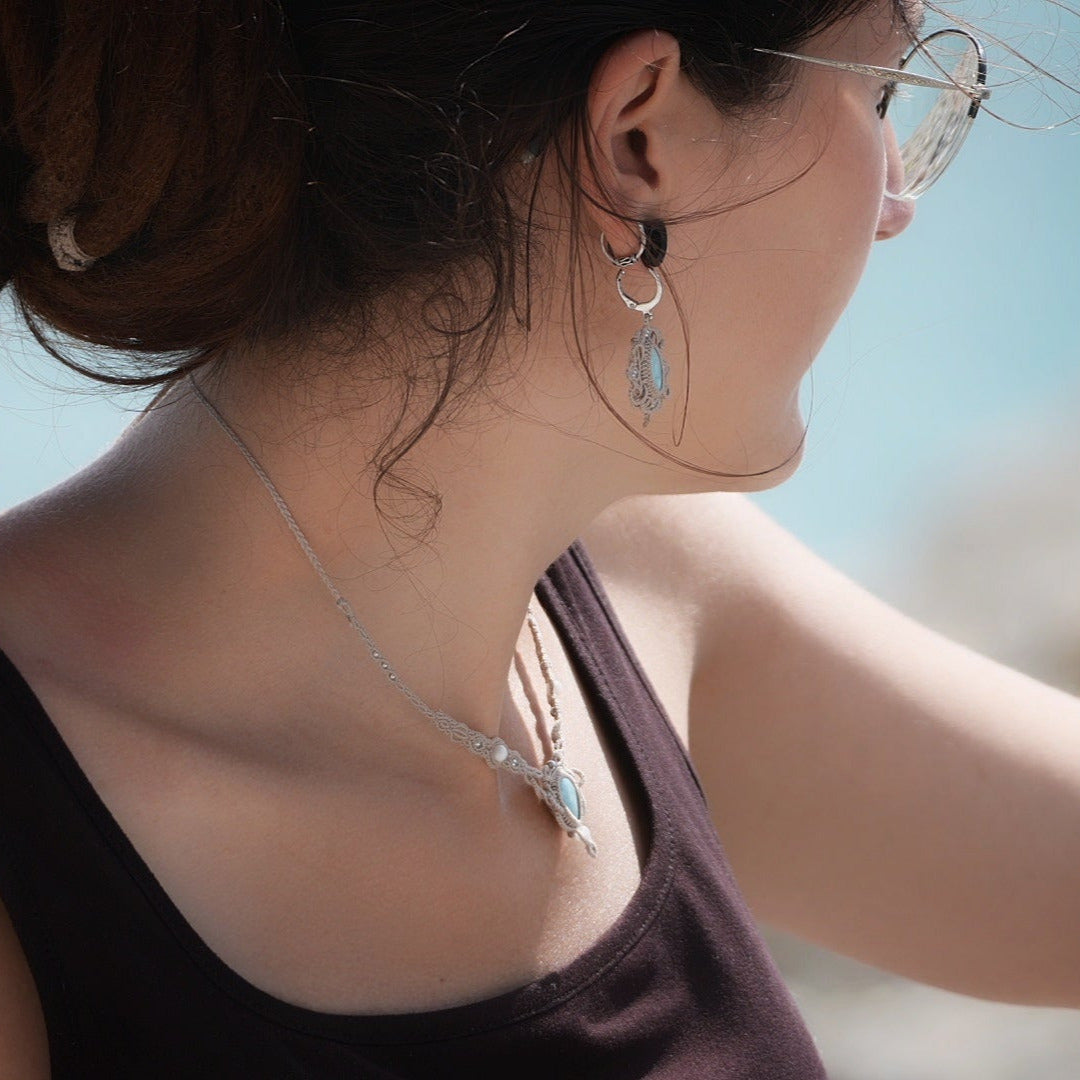 Parure Trésors d'Ecume composée d'une paire de boucles d'oreilles pendentes avec collier ras de cou assorti. Larimar bleu ciel ovales pour les boucles et en poire pointe en bas pour le collier. 3 Perles en nacre blanche pour le collier. Perles acier inoxydable argenté pour les deux pour le côté luxe et précieux. Dormeuses en acier inoxydable pour un port assuré. Fil de Coton couleur lin naturel (beige gris) de 0, 5 mm tissé en micro-macramé en relief version celtique. Bijoux porté femme brune peau caramel 
