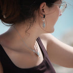 Parure Trésors d'Ecume composée d'une paire de boucles d'oreilles pendentes avec collier ras de cou assorti. Larimar bleu ciel ovales pour les boucles et en poire pointe en bas pour le collier. 3 Perles en nacre blanche pour le collier. Perles acier inoxydable argenté pour les deux pour le côté luxe et précieux. Dormeuses en acier inoxydable pour un port assuré. Fil de Coton couleur lin naturel (beige gris) de 0, 5 mm tissé en micro-macramé en relief version celtique. Bijoux porté femme brune peau caramel 