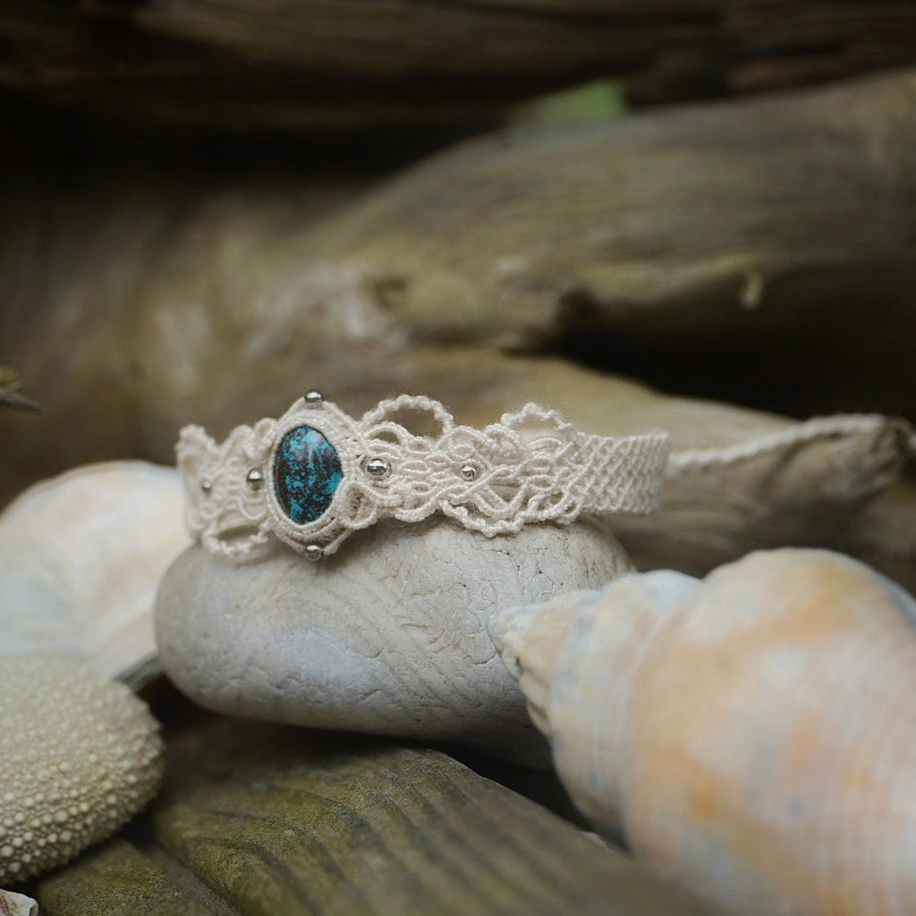 Bracelet large asymétrique. Bijou dentelle ambiance bohème hippie. Bracelet tissé en micro macramé avec fil de coton écru de 0.5 mm avec une pierre ovale de chrysocolle couleur bleu turquoise et marron foncé chocolat. Présence de part et d'autre de façon symétrique des perles en acier inoxydable argentées. Système de réglage à coulisse tubulaire à l'arrière. Design en relief et en torsade entrelacées celtiques. Ambiance plages, festival d'été et mariage boho. Bijou posé sur un galet beige et coquillages.