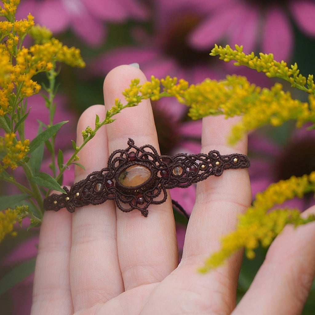 Bijou elfique en micro macramé brun avec citrine jaune et grenat rouge, posé au cœur de la nature parmi feuilles et bois. Bracelet réglable bohème de la collection Clairière de Miel, inspiré des forêts enchantées.