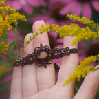 Bijou elfique en micro macramé brun avec citrine jaune et grenat rouge, posé au cœur de la nature parmi feuilles et bois. Bracelet réglable bohème de la collection Clairière de Miel, inspiré des forêts enchantées.