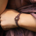 Bracelet large elfique en micro macramé brun, orné d’une citrine jaune pâle et de perles de grenat rouge rosé. Bijou bohème et artisanal réglable, porté au poignet d’une femme à la peau claire.