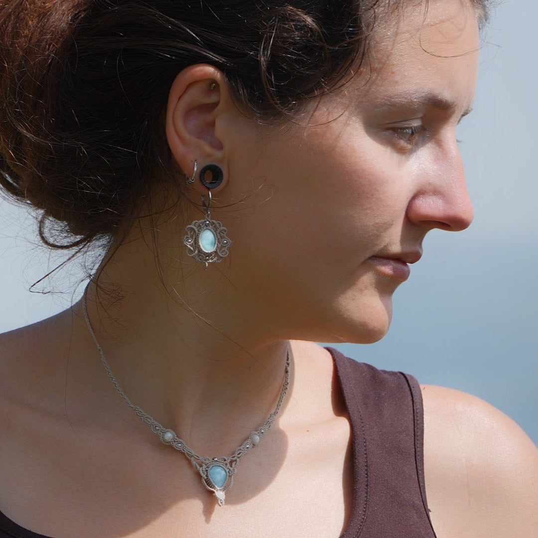 Parure Trésors d'Ecume composée d'une paire de boucles d'oreilles pendentes avec collier ras de cou assorti. Larimar bleu ciel ovales pour les boucles et en poire pointe en bas pour le collier. 3 Perles en nacre blanche pour le collier. Perles acier inoxydable argenté pour les deux pour le côté luxe et précieux. Dormeuses en acier inoxydable pour un port assuré. Fil de Coton couleur lin naturel (beige gris) de 0, 5 mm tissé en micro-macramé en relief version celtique. Bijoux porté femme brune peau caramel 